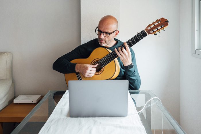 attractive middle age man, bald with glasses, playing an acoustic guitar in front of his laptop - music