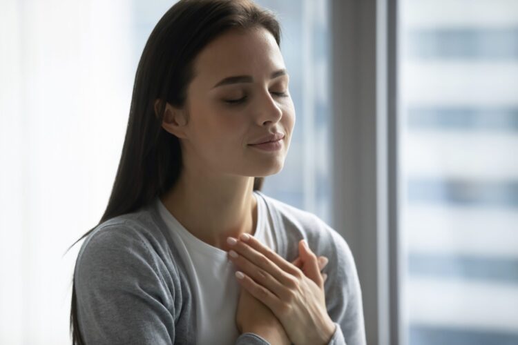 Happy millennial girl hold hands at heart chest feel thankful pray to God at workplace, calm peaceful young woman believer grateful for luck or fate, faith, superstition, gratitude concept